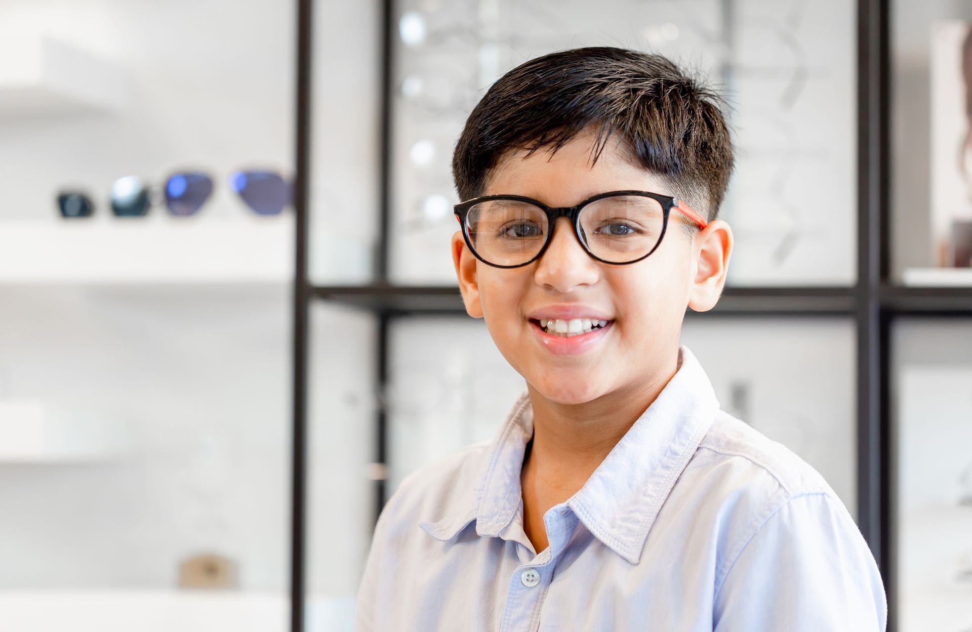 Smiling person wearing glasses and a light blue shirt in a bright room with shelves in the background