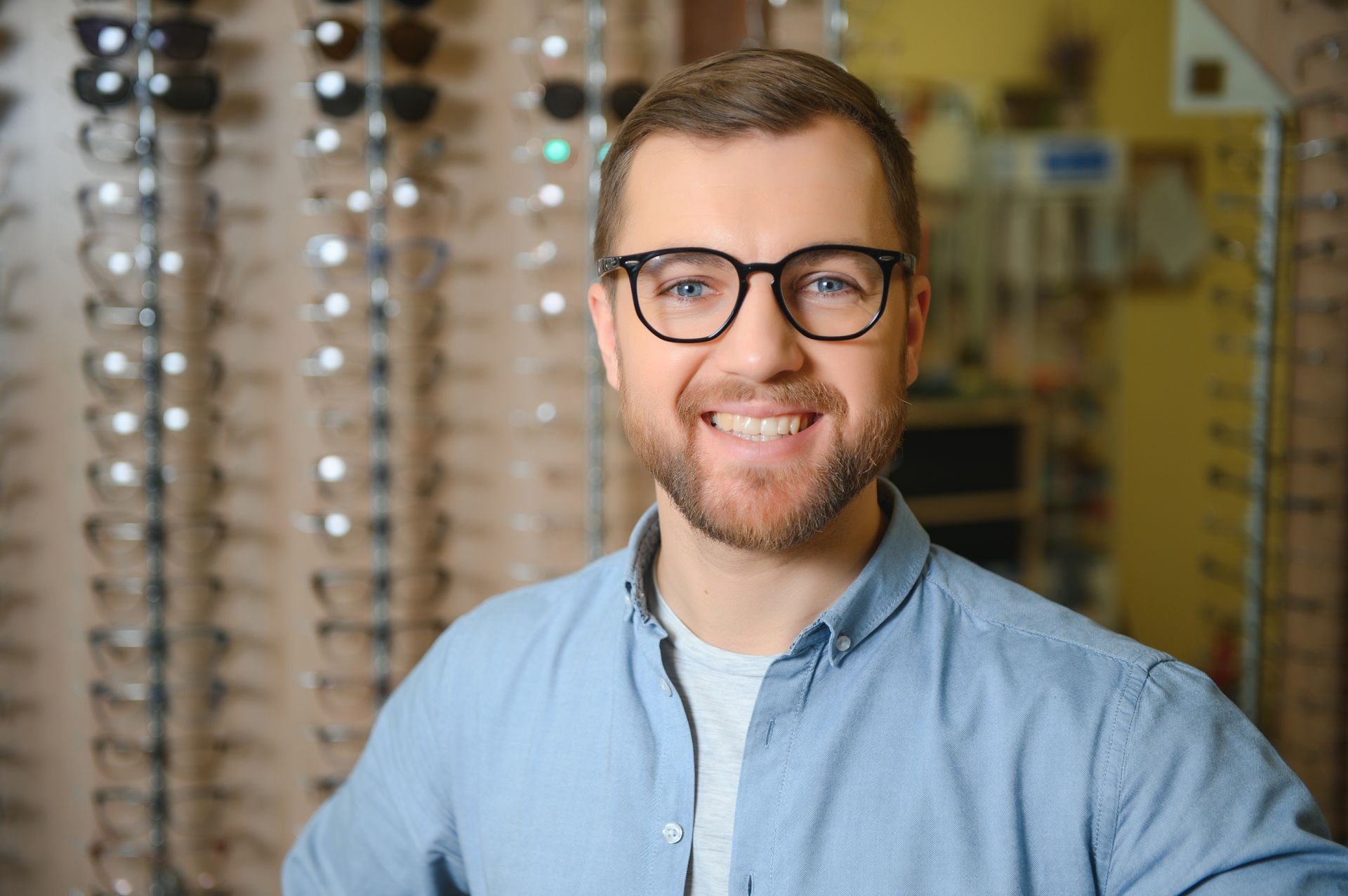 Smiling person in glasses and blue shirt in an optical shop with eyeglass frames behind them
