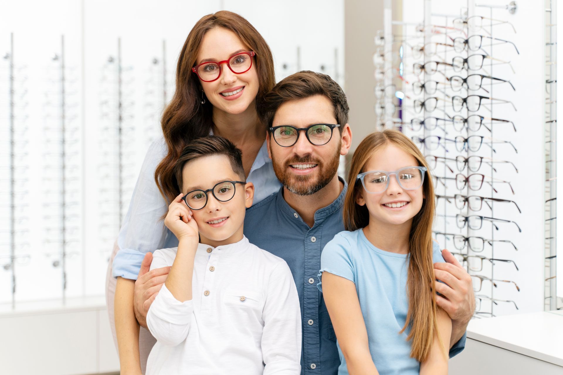Family smiling at an eyeglasses shop, with two adults and two children wearing glasses.