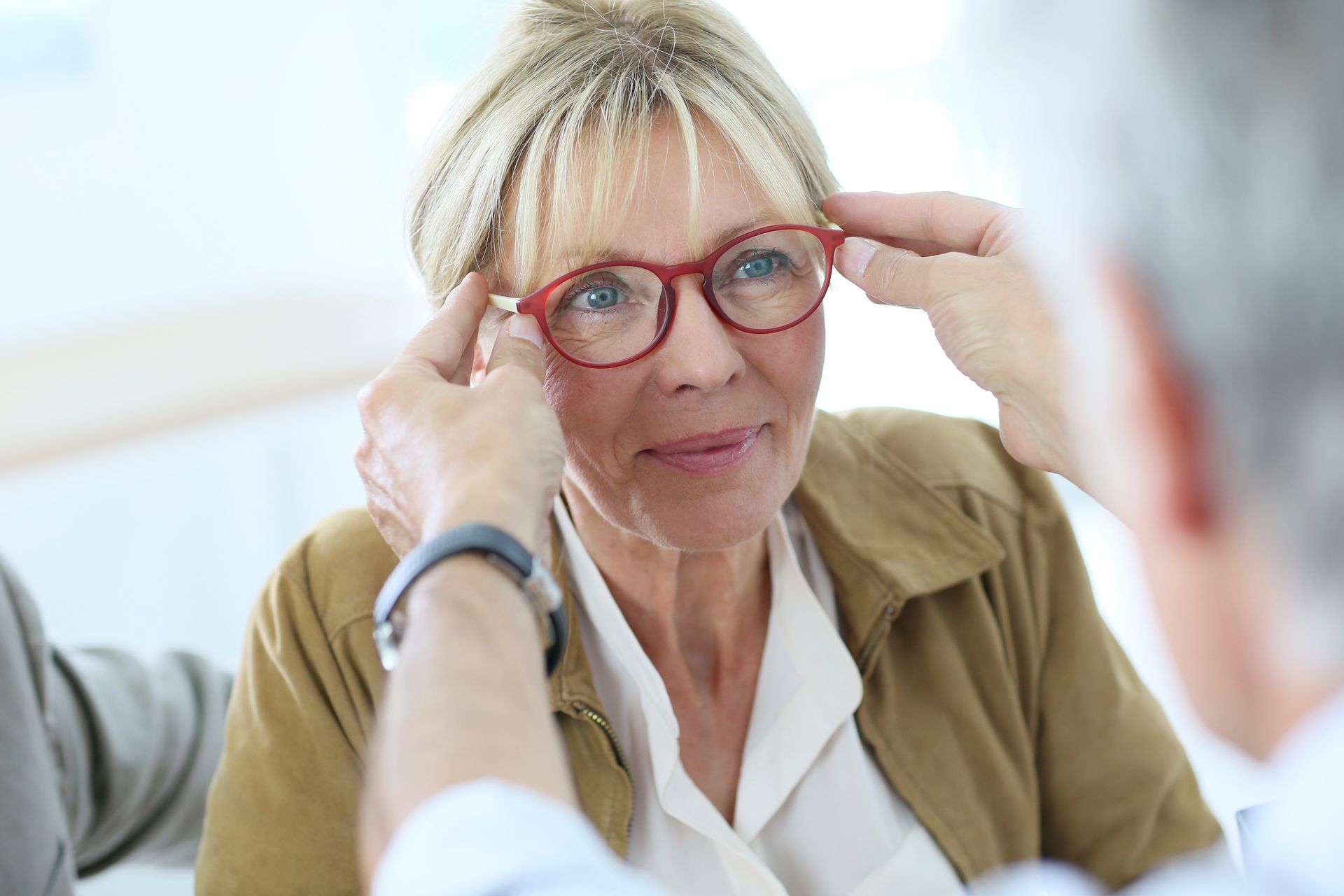 Person adjusting red glasses during an eye exam, with hands near their face in a bright clinic