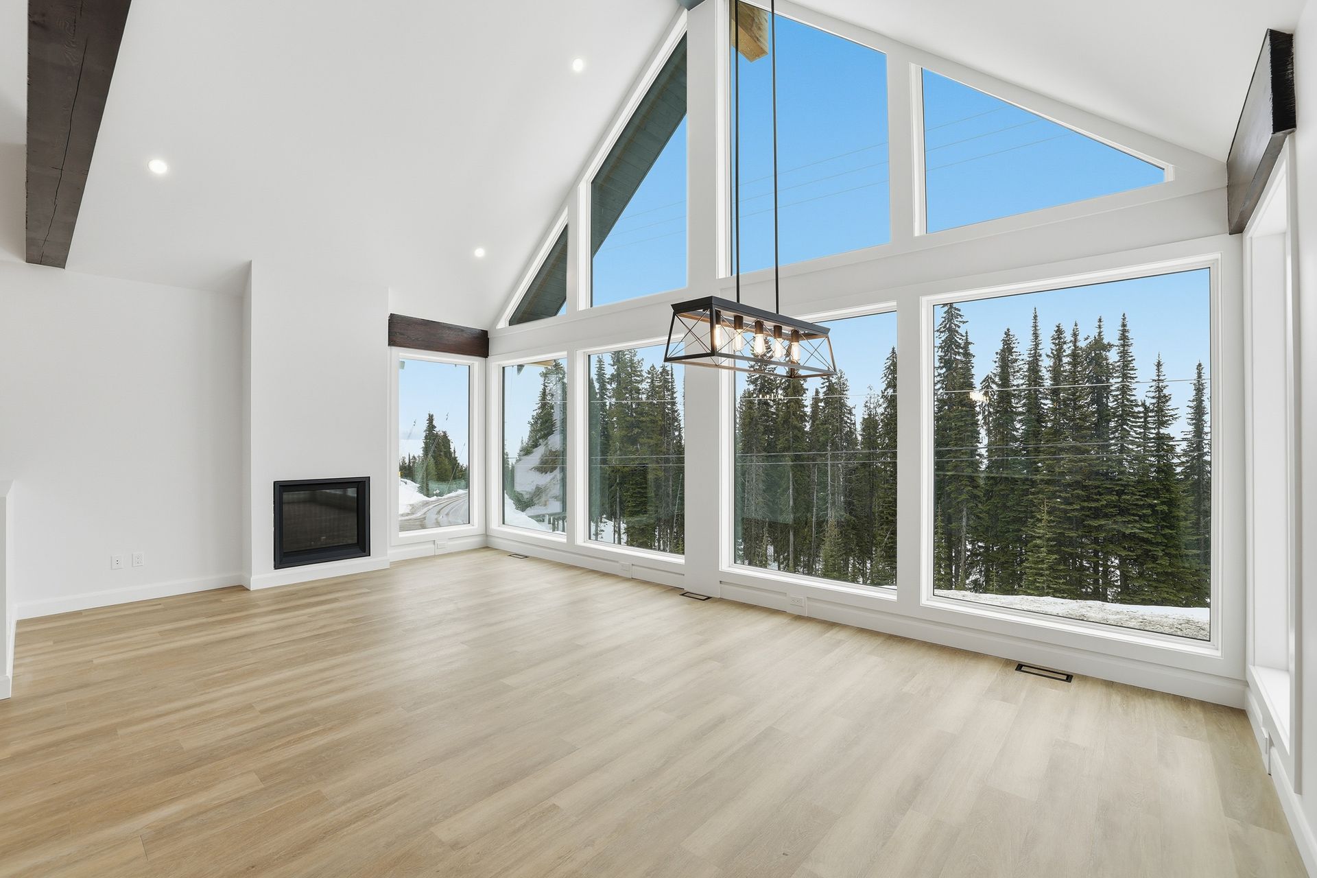 Bright modern living room with vaulted ceiling, fireplace, and floor-to-ceiling windows overlooking evergreens