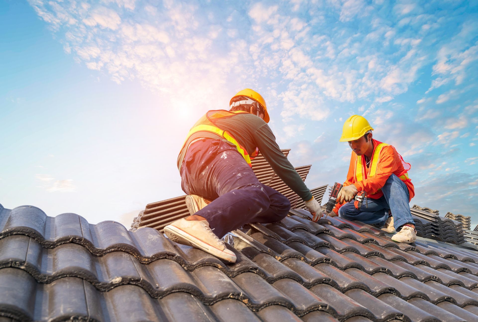 Two construction workers in orange vests and yellow helmets installing roof tiles on a sloped roof