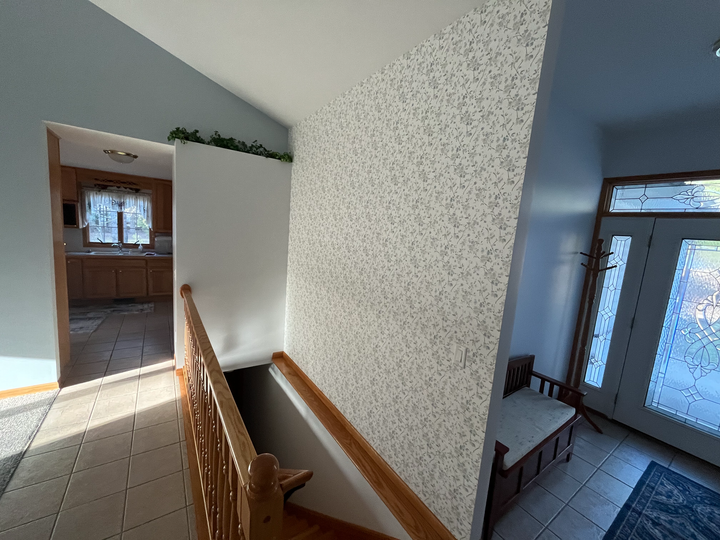 Stair landing with wooden railing, patterned wallpaper, and a hallway leading to a front door and tiled entryway