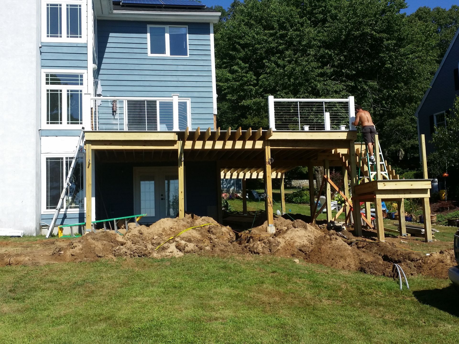 A partially constructed wooden deck on a grassy hill; person working on the upper level, surrounded by dirt piles and a house.