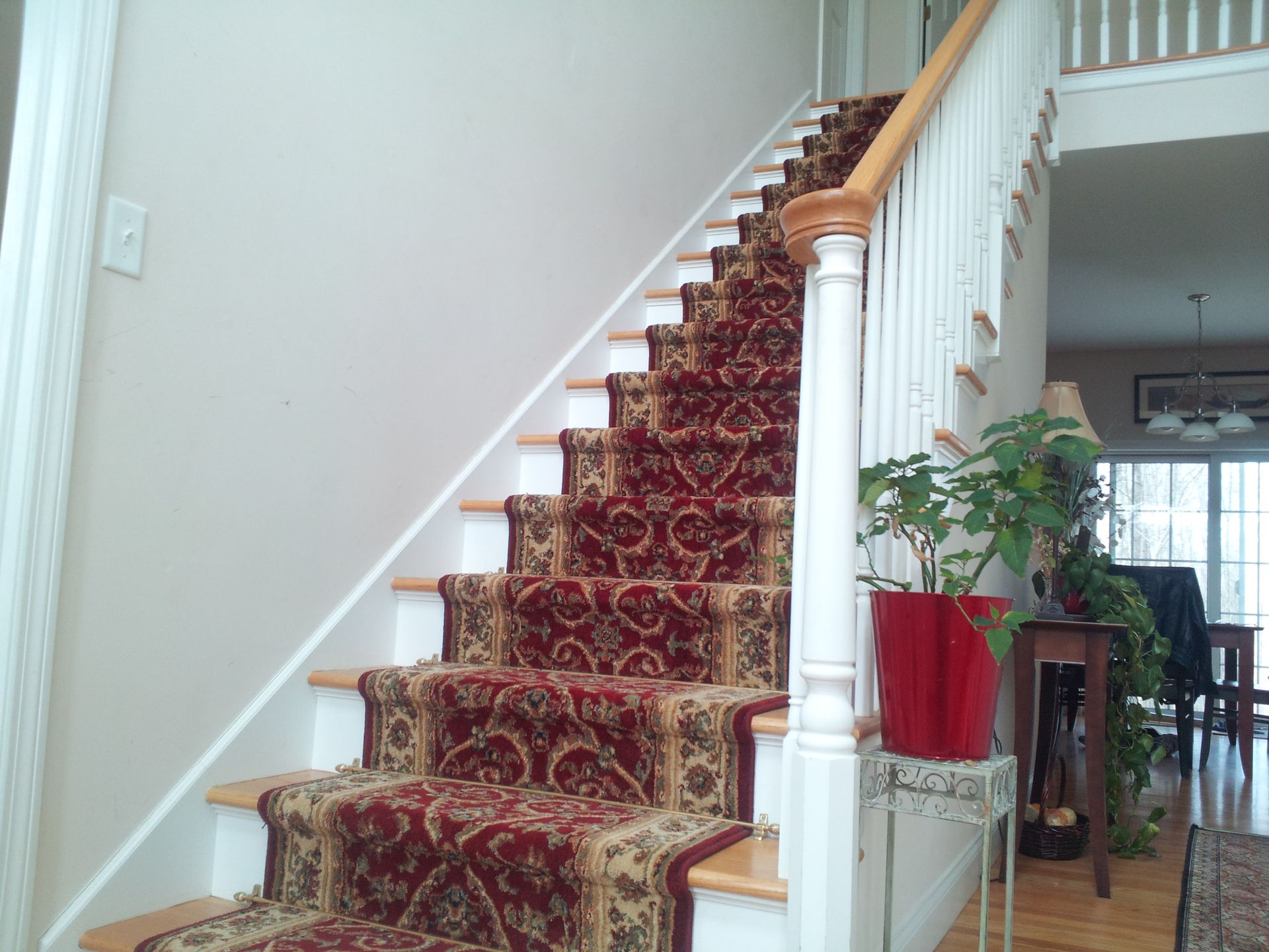 Staircase with patterned red and beige carpet, white banister, and light wood steps.