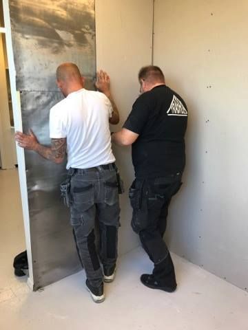 Two workers installing a metal panel in a corner. One person supports the panel. White walls, light floor.
