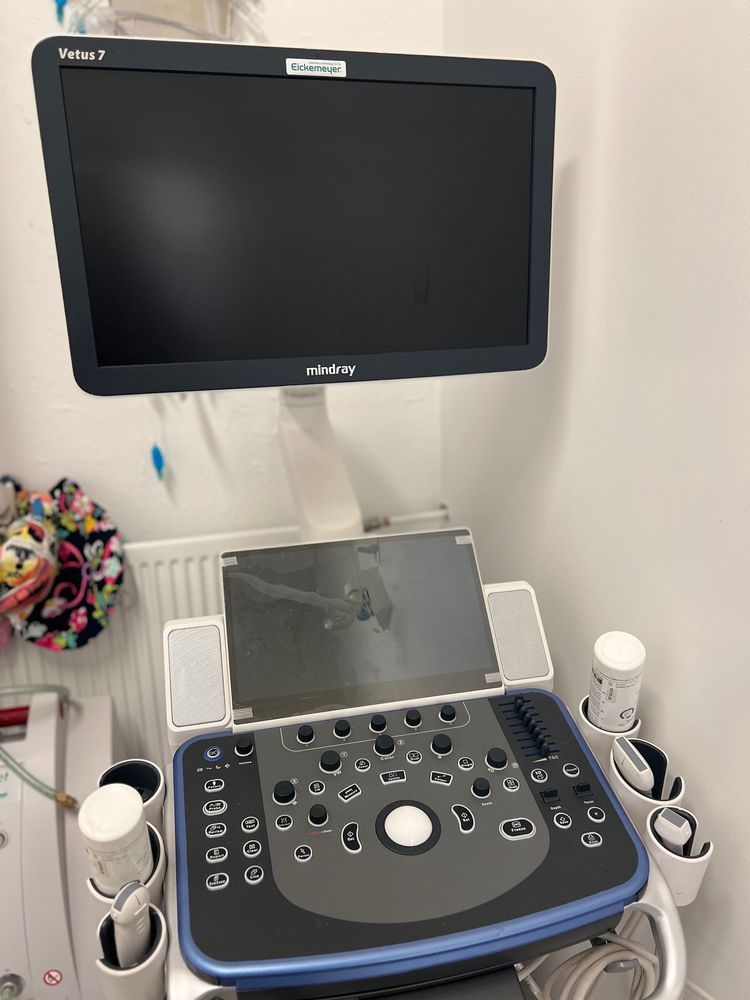 Ultrasound machine with a dark screen, control panel, and probe holders, in a medical setting.