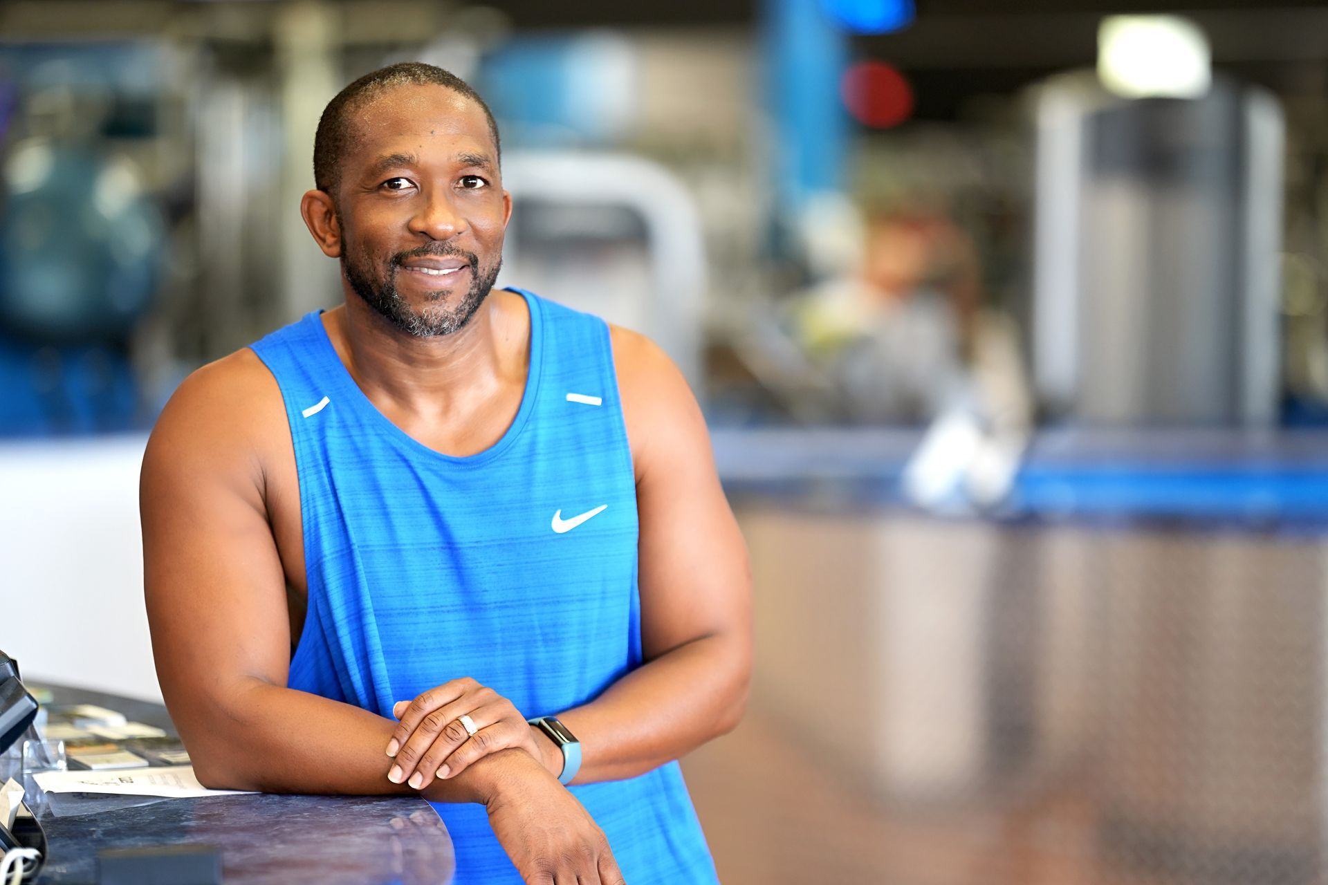 A man in a blue tank top is leaning on a counter in a gym.