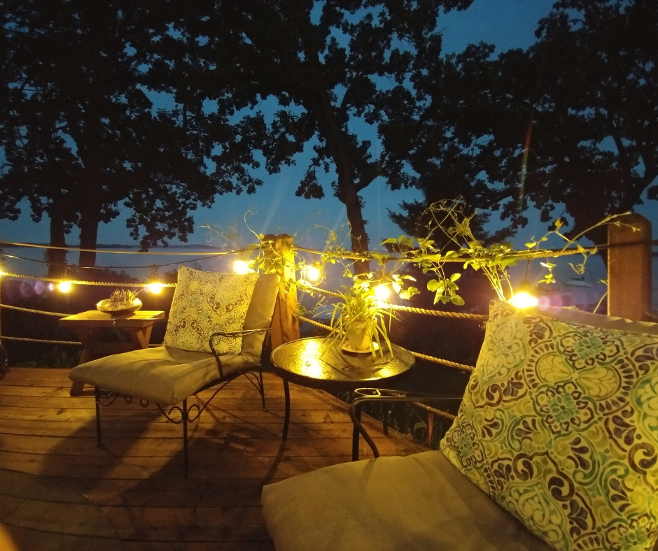 Patio deck at dusk with string lights, cushioned chairs, and a small table overlooking dark trees and water