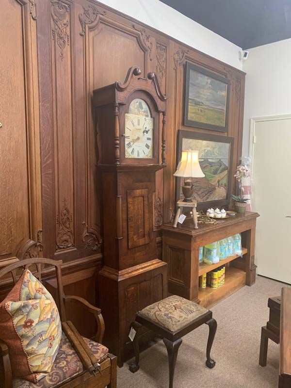 Antique wooden clock and cabinet in a cozy room with lamp, paintings, chair, and patterned stool