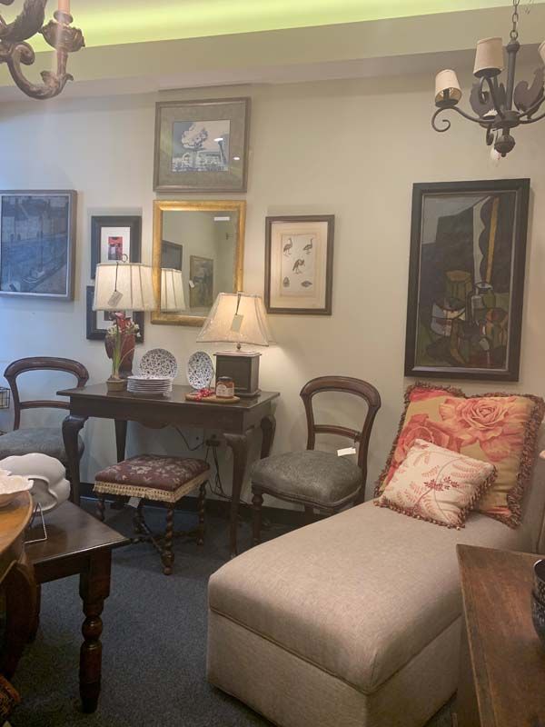Cozy living room with upholstered chairs, framed art, a lamp, and a beige ottoman under a chandelier.