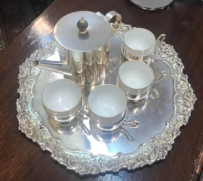 Silver tea set on a decorative tray with several covered cups on a dark table