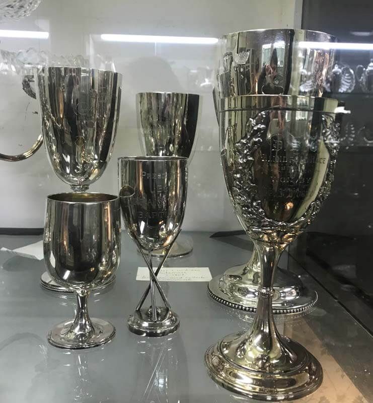 Five reflective silver trophy cups displayed on a glass shelf.