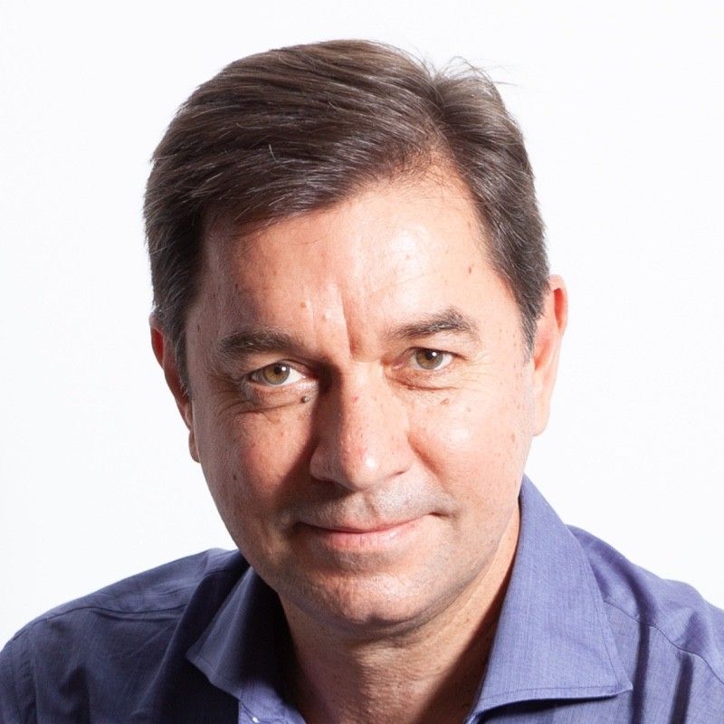 Man in a blue blazer and striped shirt, smiling, indoors with plants and light wood floors.