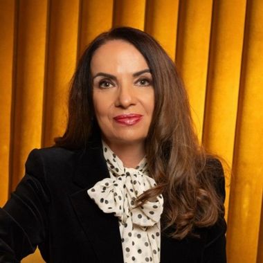 Woman with wavy brown hair, wearing a dark blazer, smiling, in front of a neutral background.
