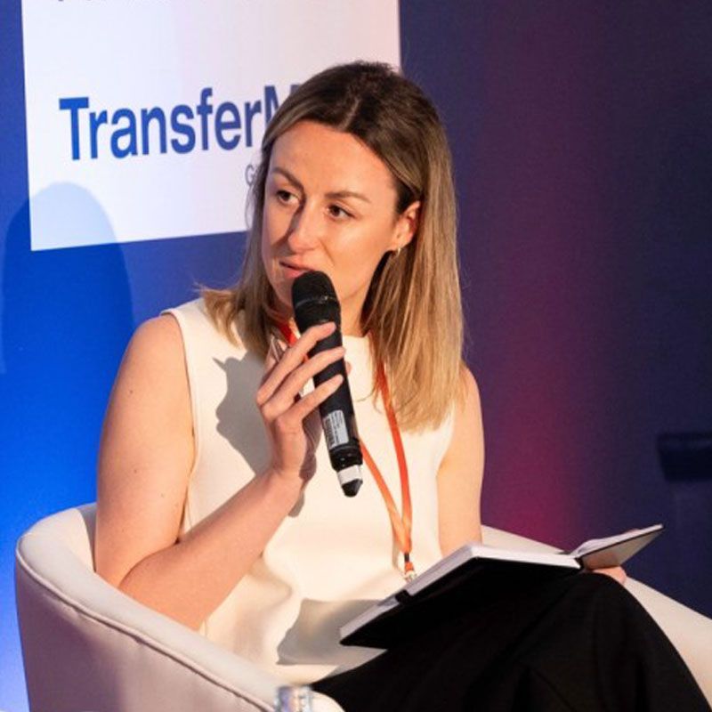 Woman speaking into a microphone, seated, taking notes at a conference.