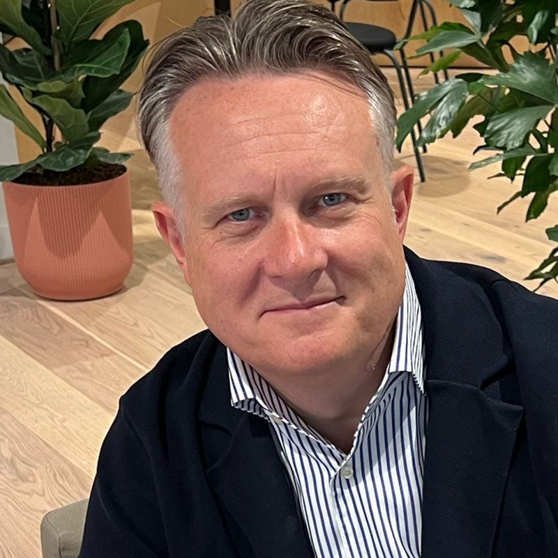 Man in a blue blazer and striped shirt, smiling, indoors with plants and light wood floors.