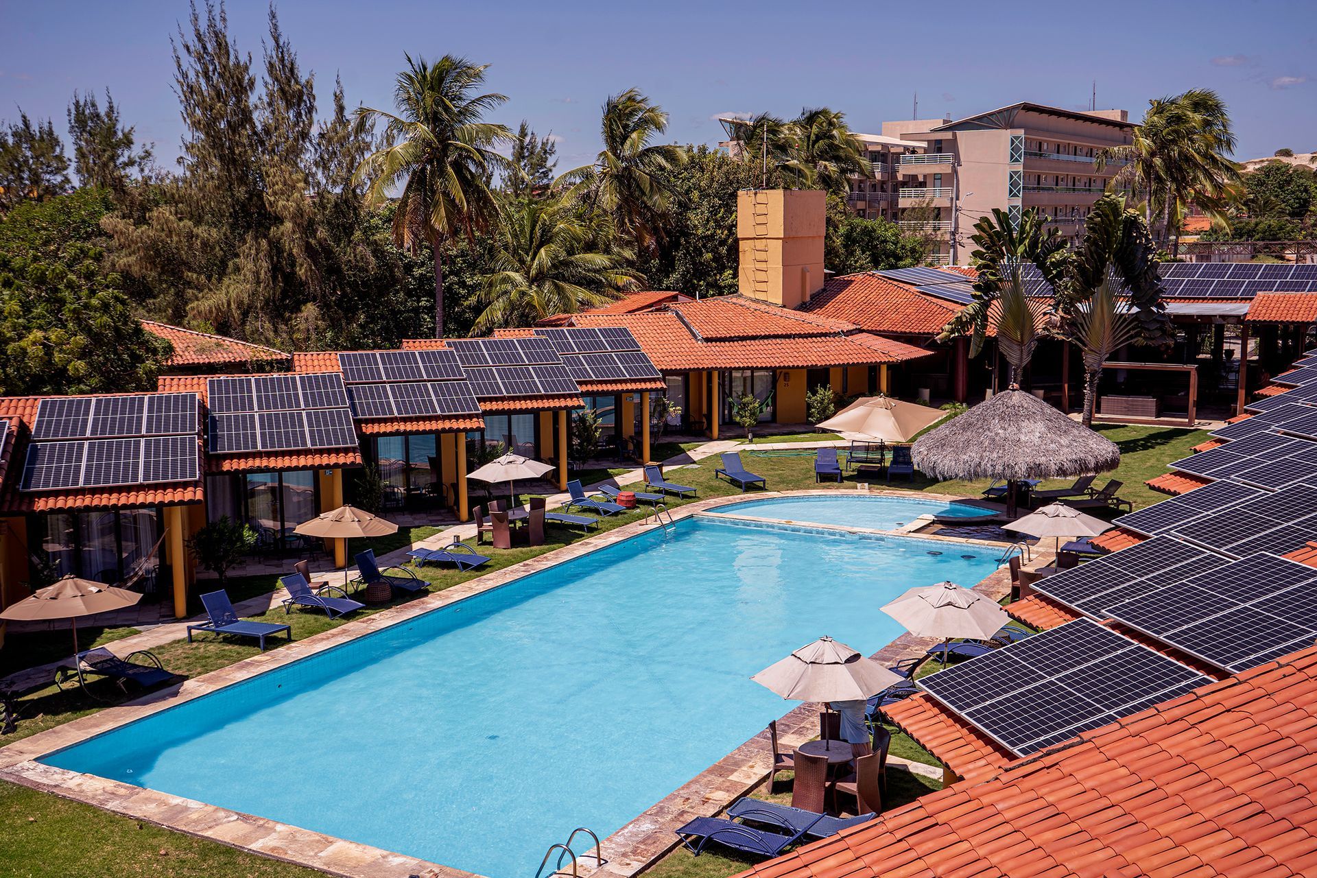 Piscina do resort rodeada por telhados de telha vermelha, palmeiras e espreguiçadeiras sob um céu ensolarado.