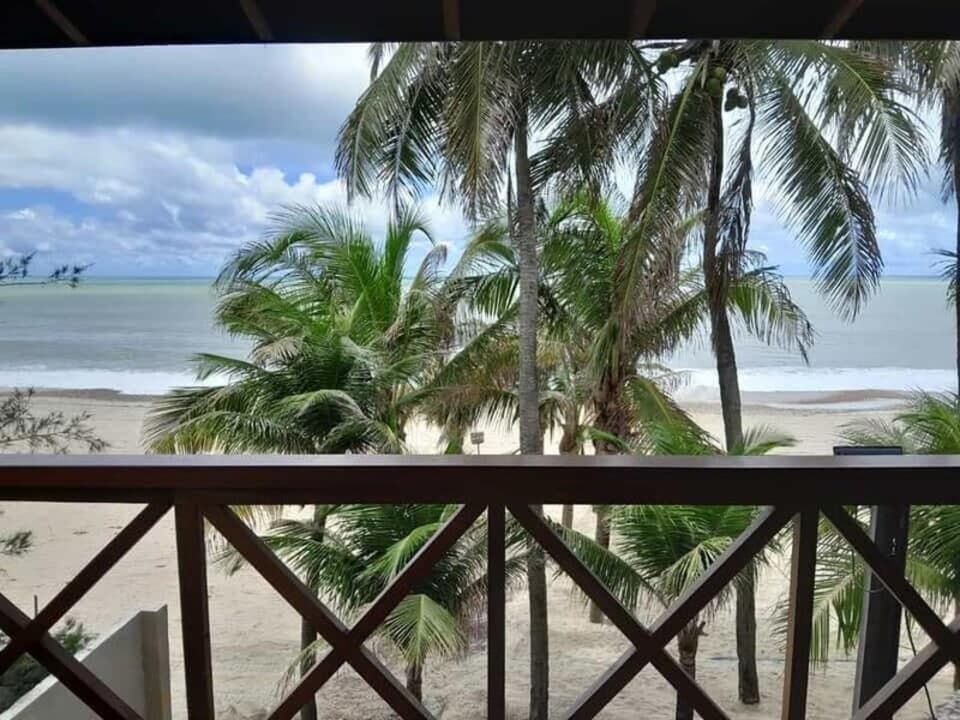 Vista de uma praia tropical com palmeiras, oceano calmo e céu nublado, a partir do parapeito de uma varanda de madeira.