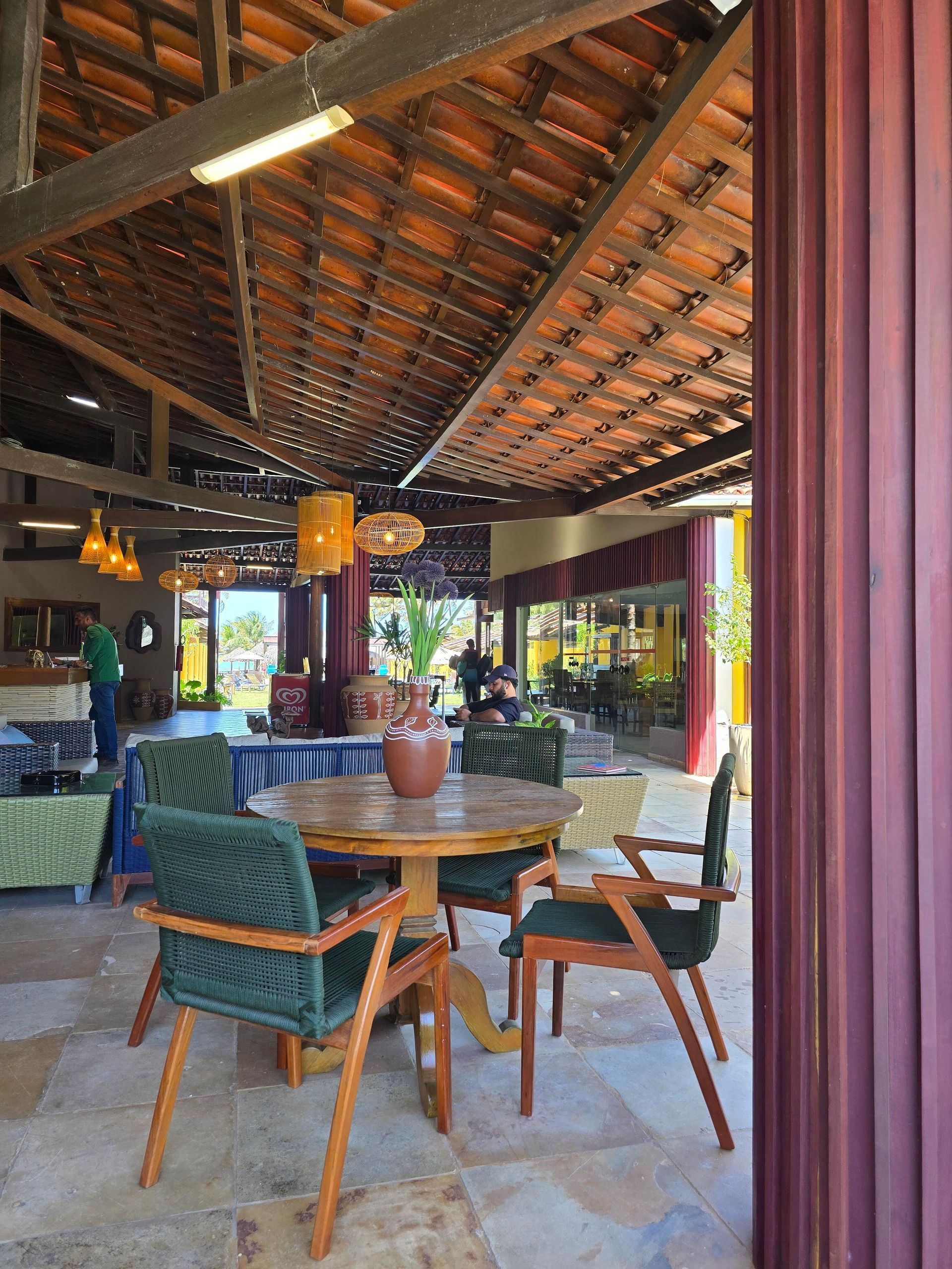 Restaurante ao ar livre com mesas e cadeiras de madeira sob um telhado de telhas, com vista para a vegetação.