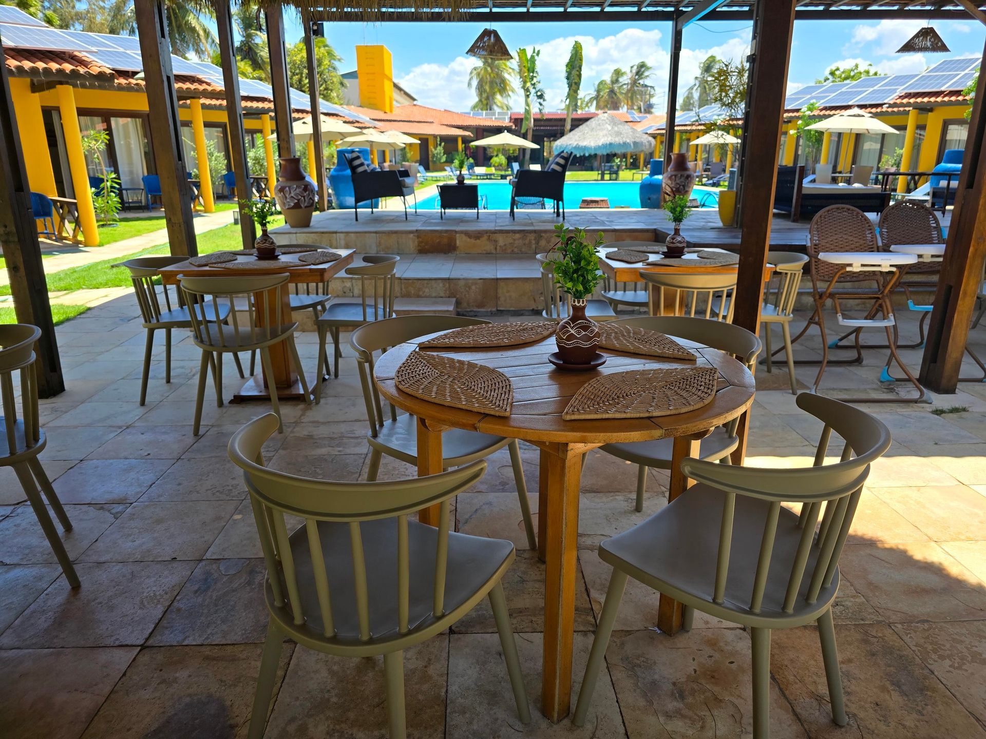 Café ao ar livre com mesas e cadeiras de madeira, com vista para uma piscina iluminada e para a praia.