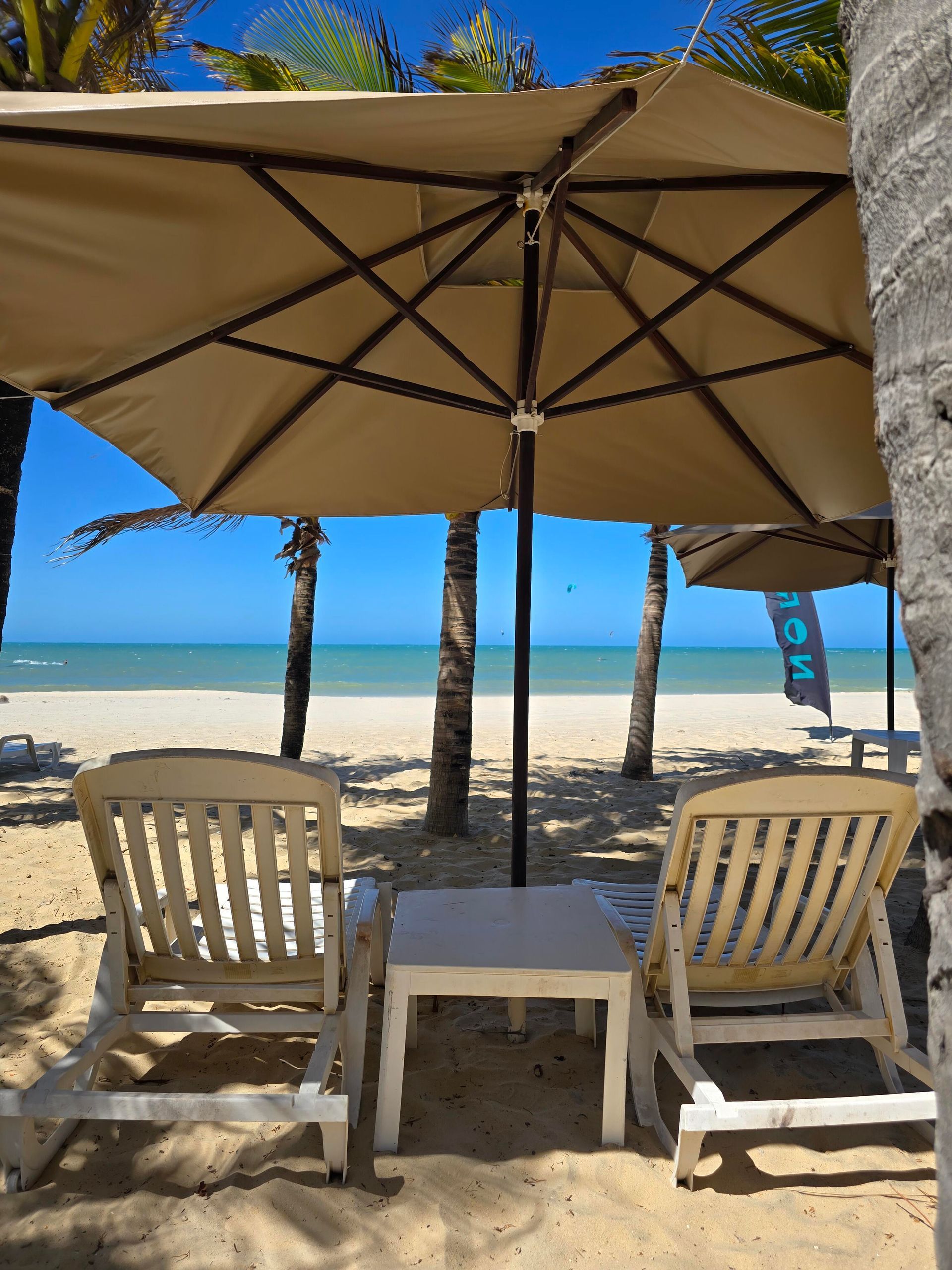 Duas cadeiras de praia e uma pequena mesa sob um guarda-sol bege em uma praia tropical ensolarada.