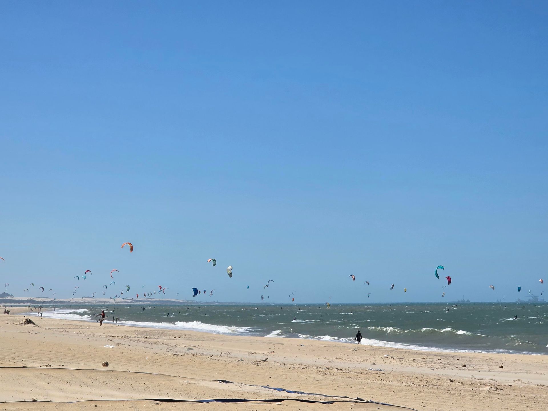 Praia ensolarada com ondas e várias pipas coloridas voando sobre o oceano.