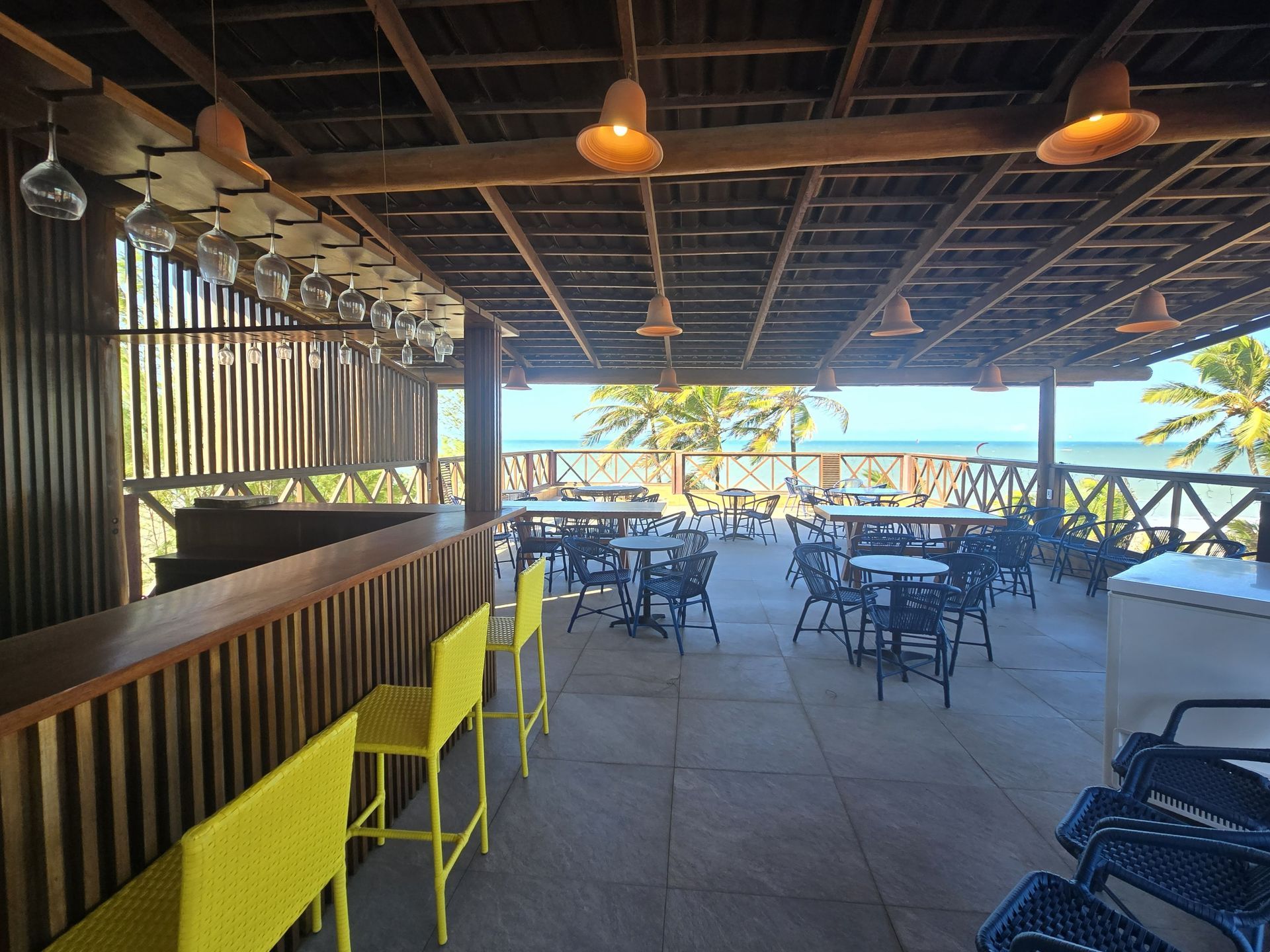Pátio de restaurante ao ar livre com banquetas amarelas, mesas escuras e vista para o mar através de um parapeito de madeira.