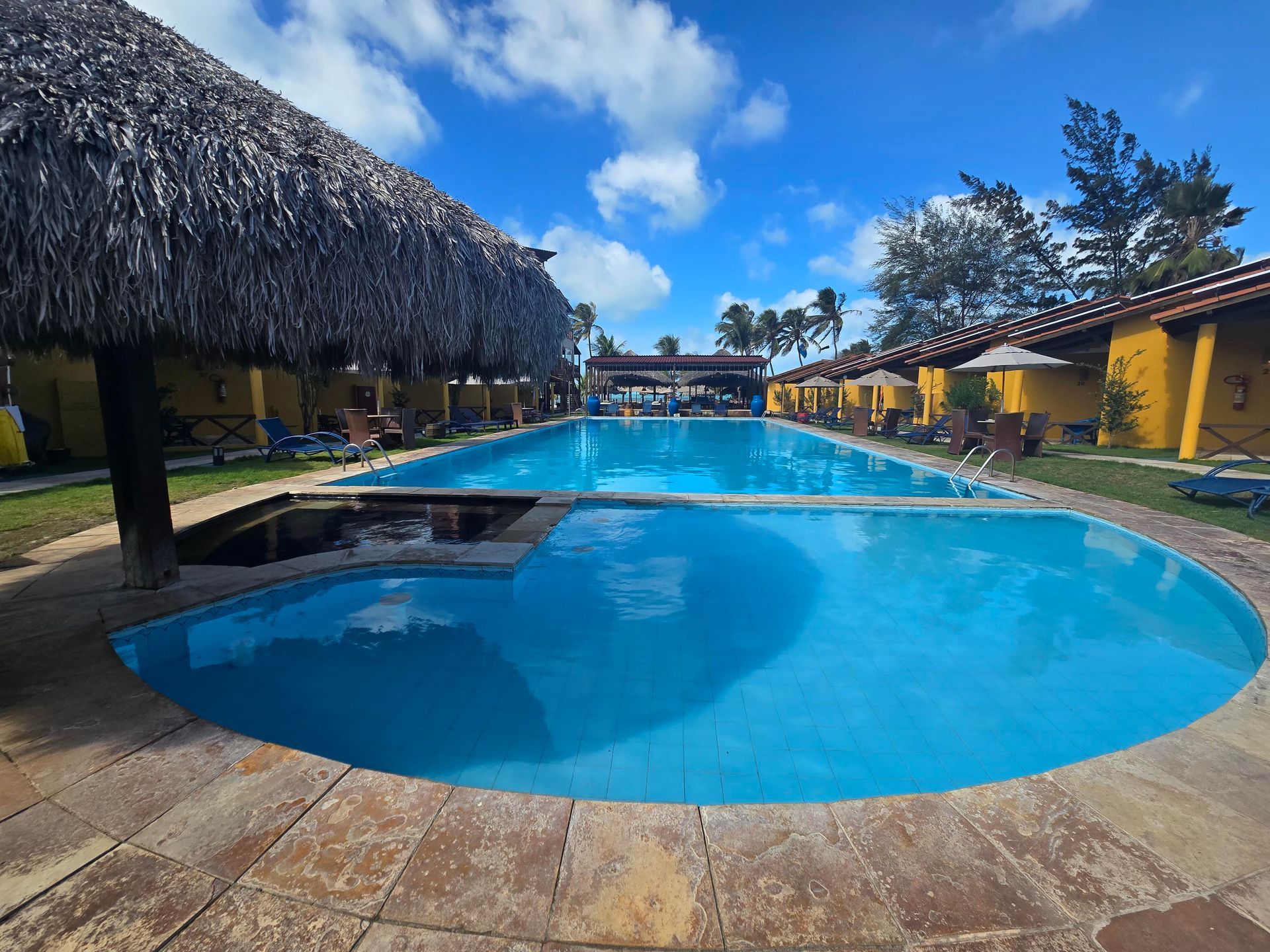 Piscina de resort com água azul cristalina, coberturas de palha para sombra e cabanas amarelas sob um céu claro.