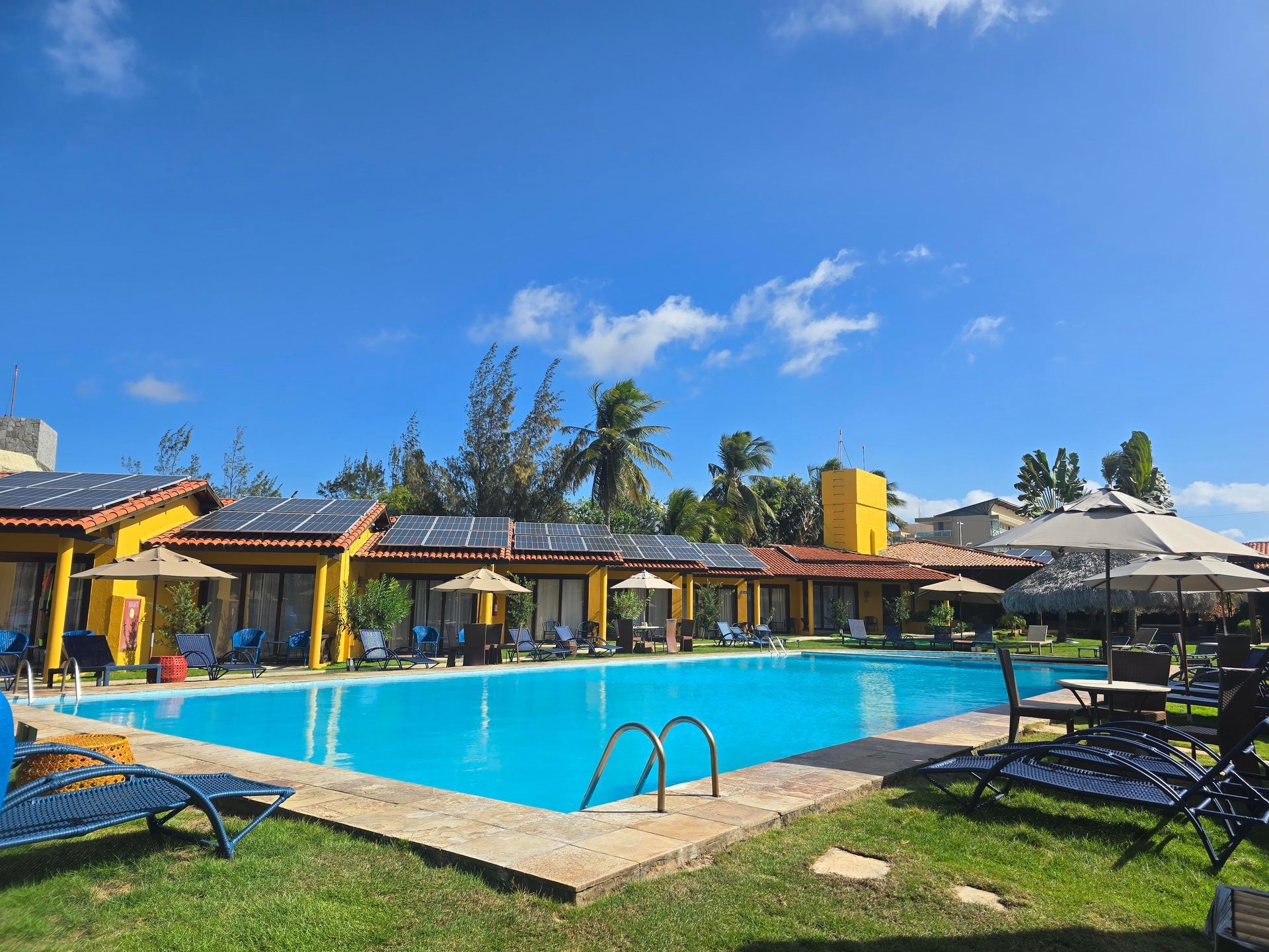 Piscina do resort com espreguiçadeiras, guarda-sóis e prédios amarelos sob um céu azul brilhante.