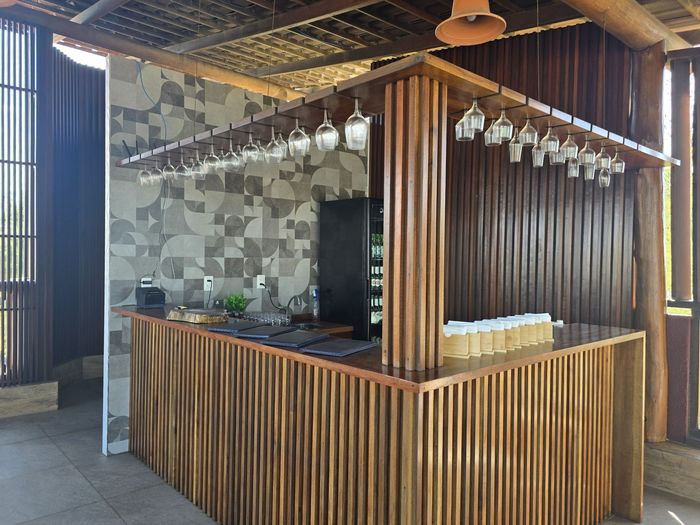 Quiosque de café com balcão de madeira, parede de azulejos ao fundo e luzes pendentes em um espaço interno moderno.
