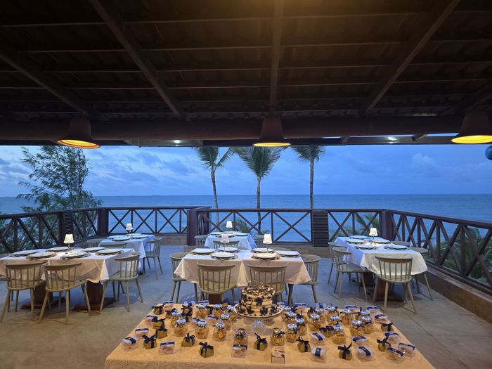 Ambiente para refeições ao ar livre à beira-mar, com mesas cobertas com toalhas brancas e vista para o oceano ao pôr do sol.