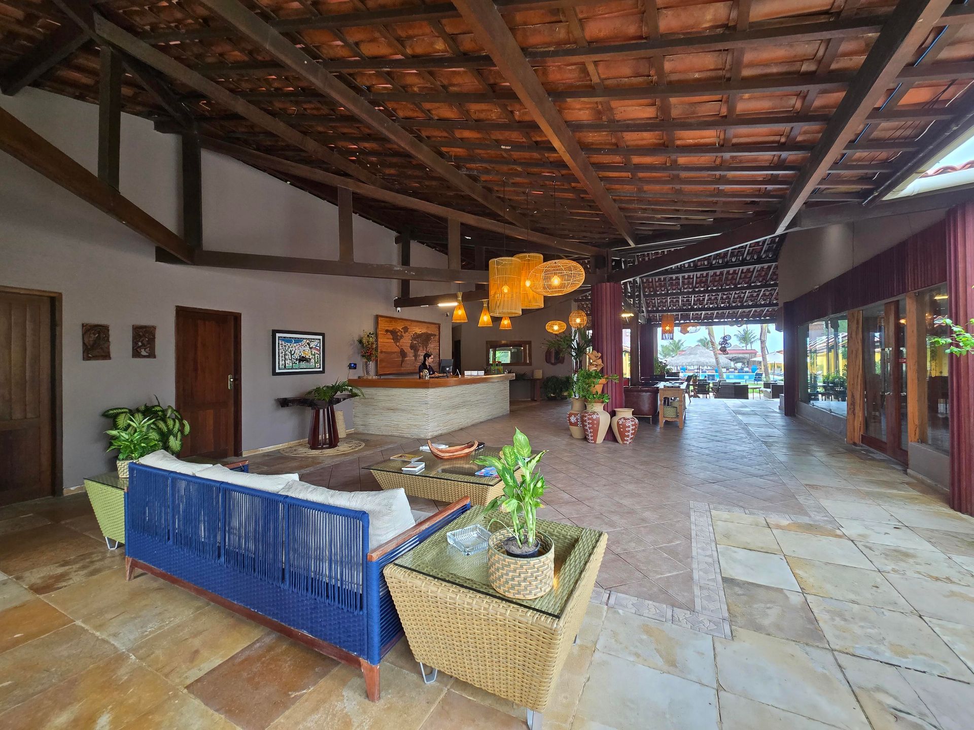 Lobby de resort ao ar livre com vigas de madeira, banco azul, balcão de recepção e plantas em vasos.