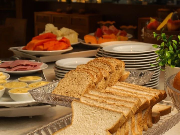 Mesa de buffet com pão fatiado, doces, frutas e pratos empilhados, prontos para servir.