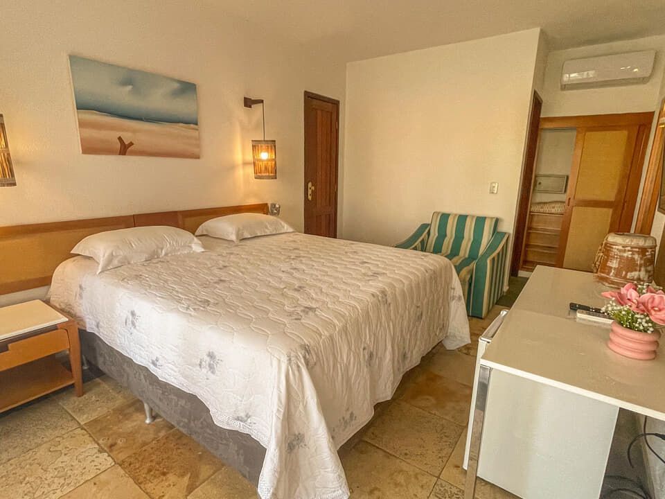 Quarto bem arrumado com duas camas de solteiro, iluminação aconchegante, piso de azulejo e uma pequena escrivaninha junto à janela.