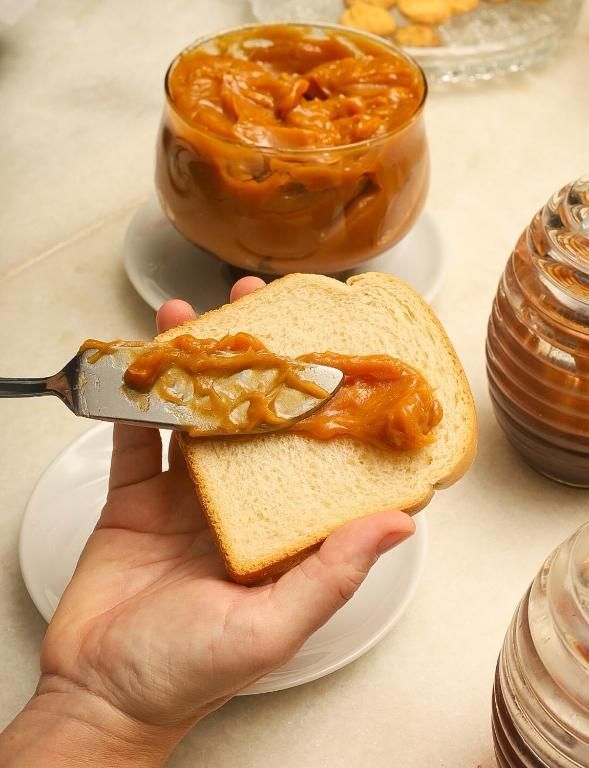 Uma pessoa espalha geleia de laranja em uma fatia de pão branco, com potes de geleia em uma mesa atrás.