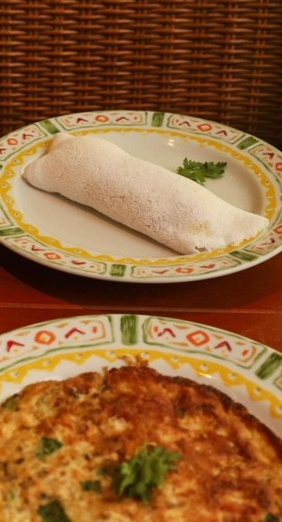 Dois pratos servidos individualmente: um dosa branco enrolado com chutney e uma omelete dourada, ambos decorados com ervas.