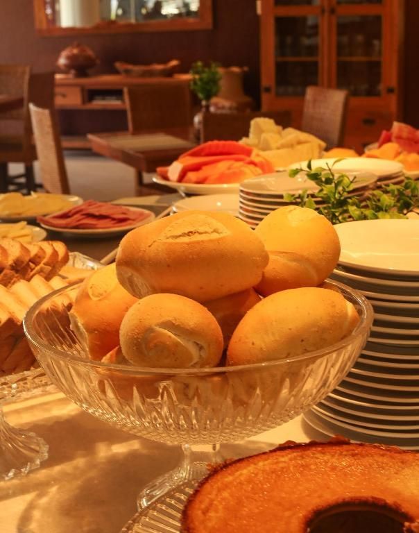 Pãezinhos em uma tigela de vidro sobre uma mesa de jantar, com pratos, saladas e um doce de café da manhã em primeiro plano.