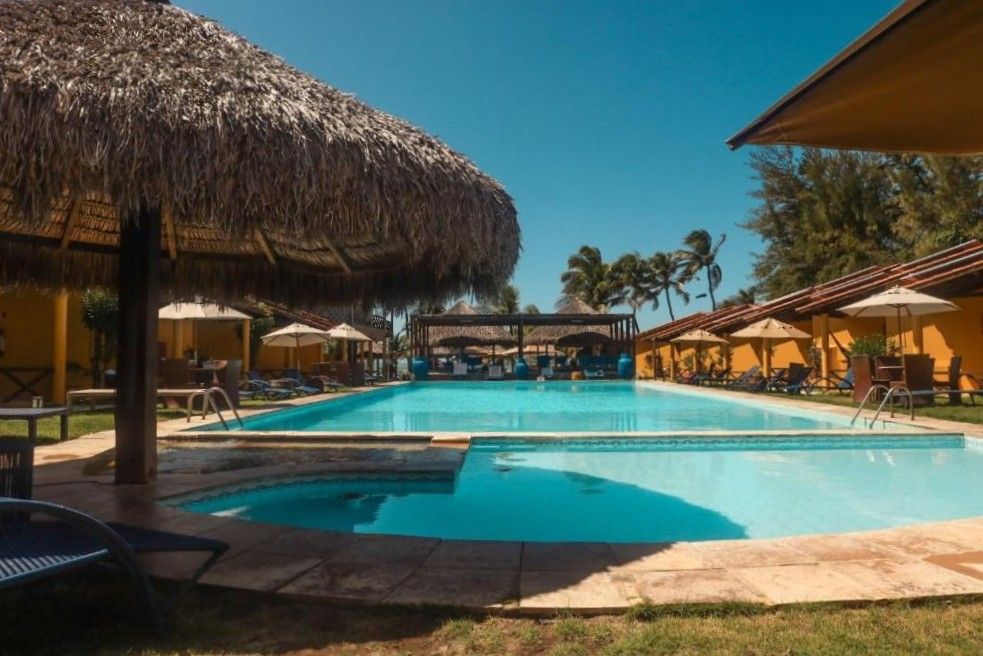 Piscina do resort com guarda-sóis de palha, espreguiçadeiras e água azul sob um céu ensolarado.