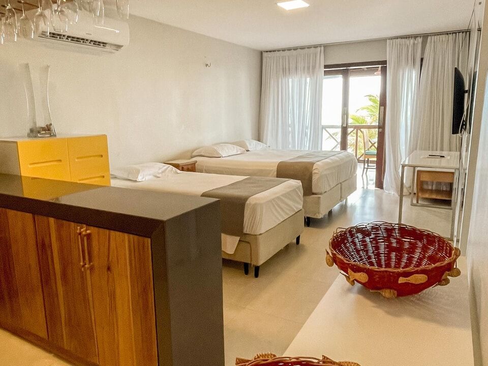 Quarto iluminado com duas camas, cortinas brancas, cômoda amarela e uma cesta vermelha no chão.