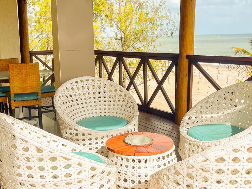 Área de estar à beira-mar com cadeiras de vime branco, almofadas turquesa e uma pequena mesa com vista para o oceano.