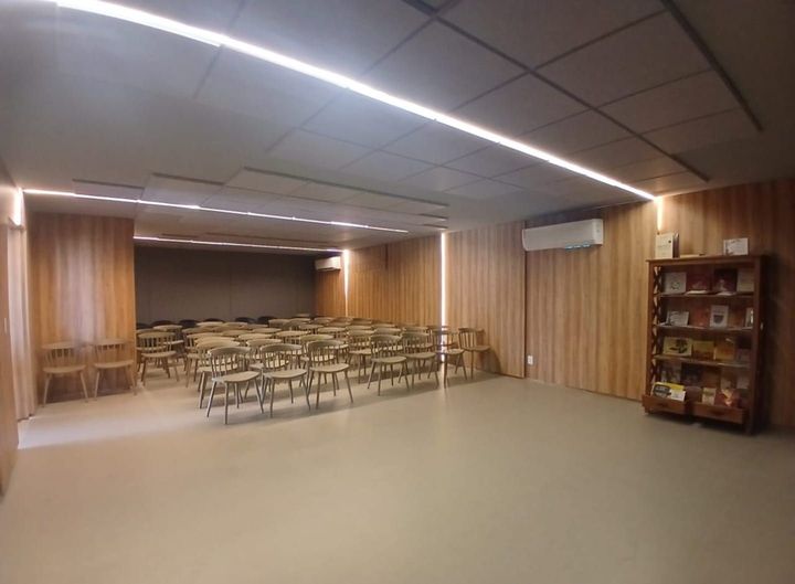 Sala de reuniões vazia com fileiras de cadeiras, paredes revestidas de madeira e luzes fluorescentes no teto.