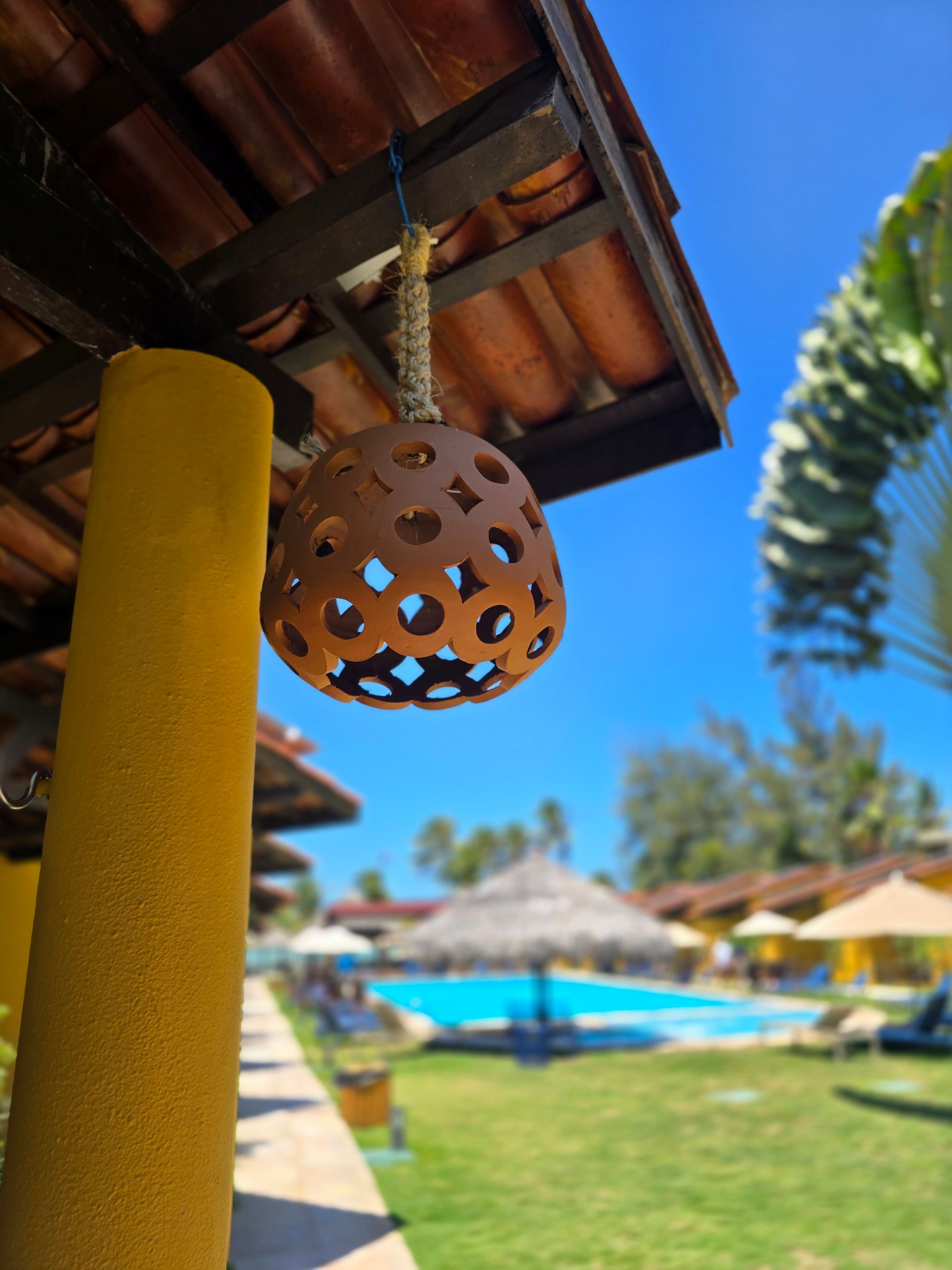 Lanterna de terracota pendurada ao lado de um poste amarelo na piscina ensolarada de um resort, com guarda-sóis e palmeiras.