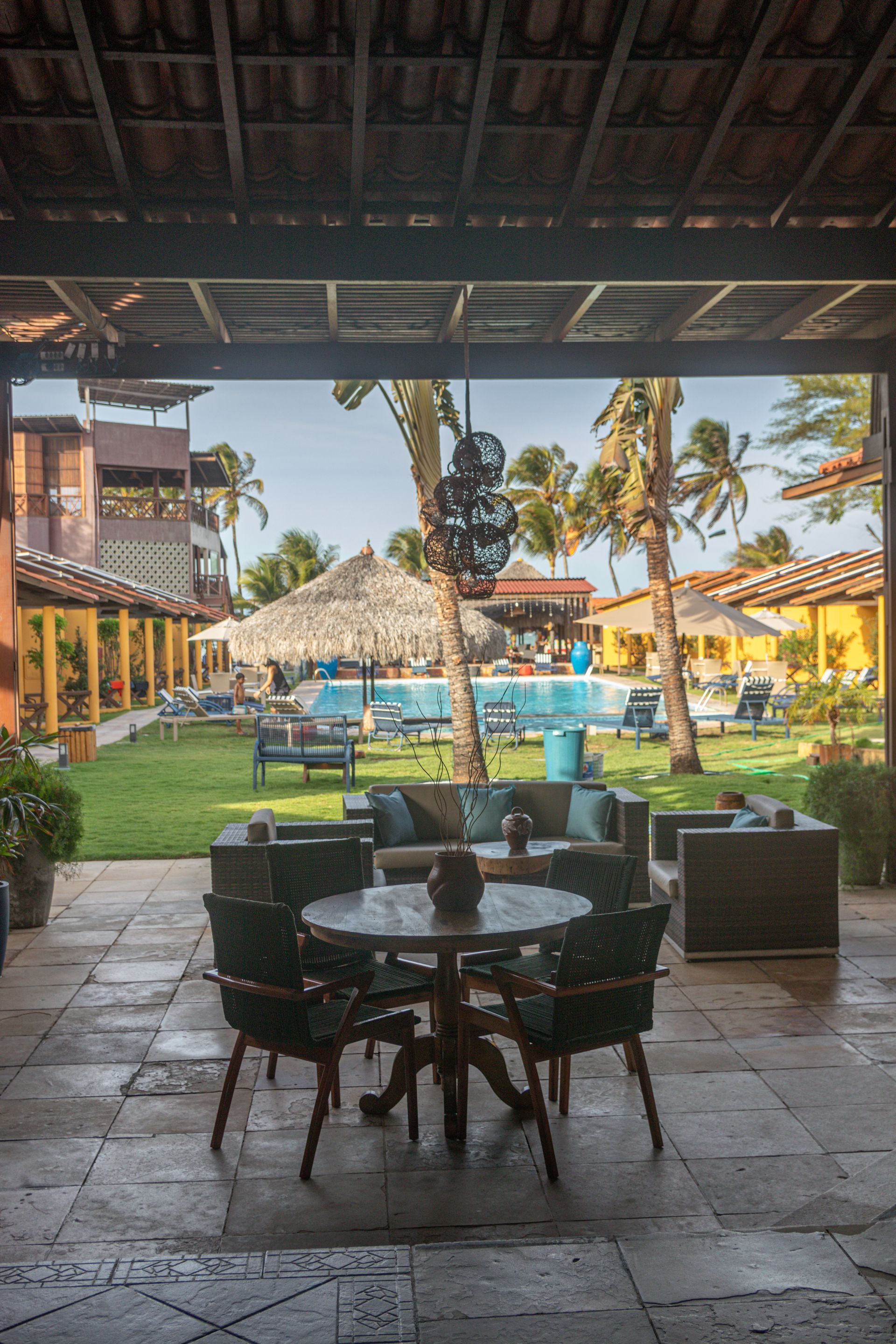 Pátio coberto com cadeiras com vista para uma piscina tropical e guarda-sóis de palha em um resort.