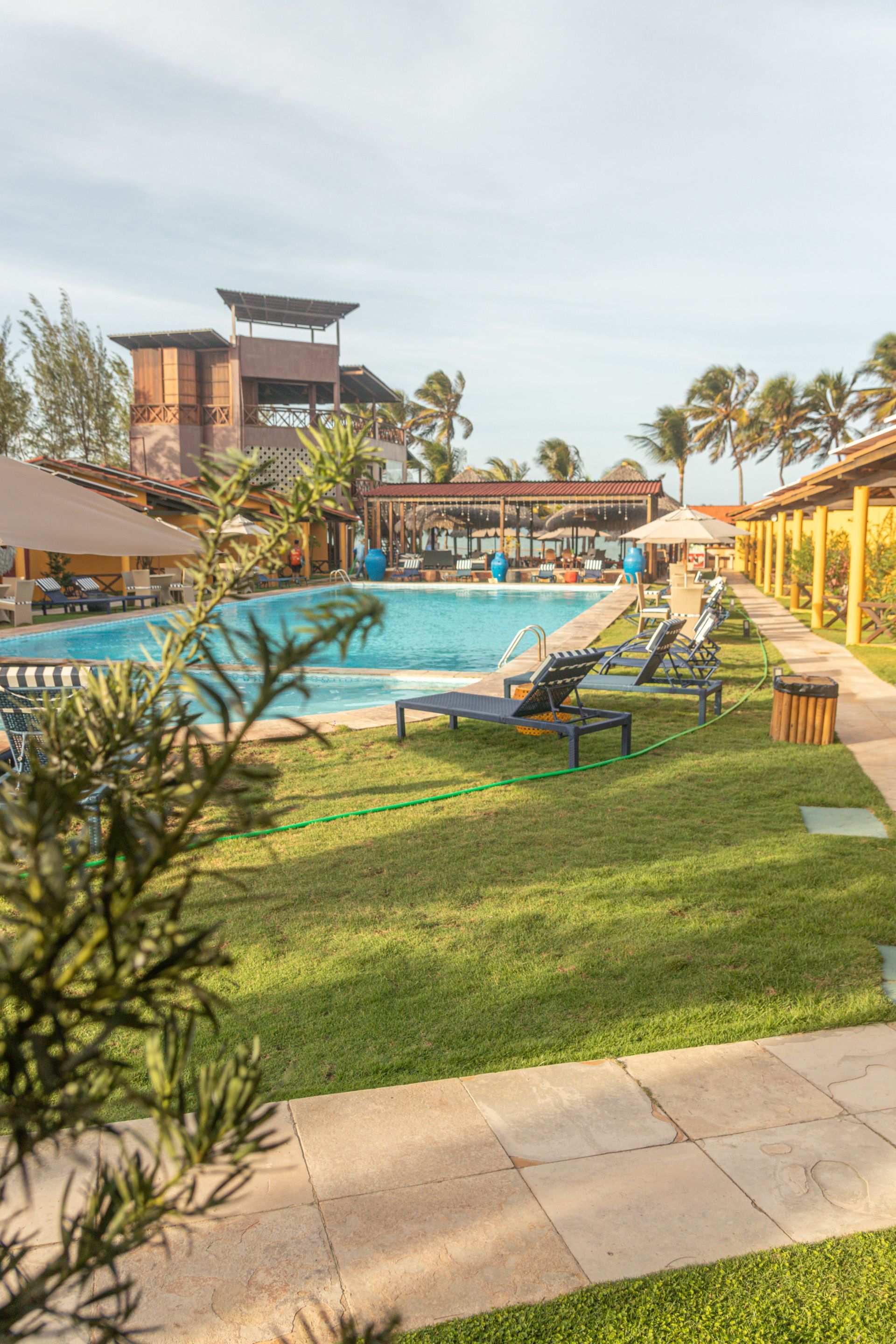 Piscina do resort com espreguiçadeiras, palmeiras e um céu azul límpido.
