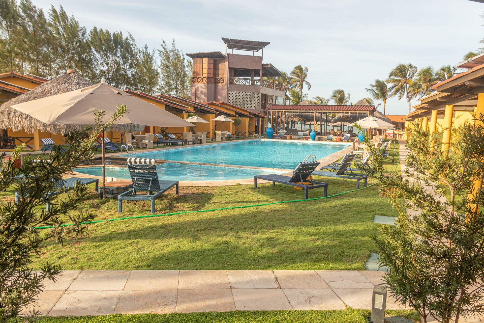 Piscina do resort com espreguiçadeiras, guarda-sóis e paisagismo tropical sob um céu ensolarado.