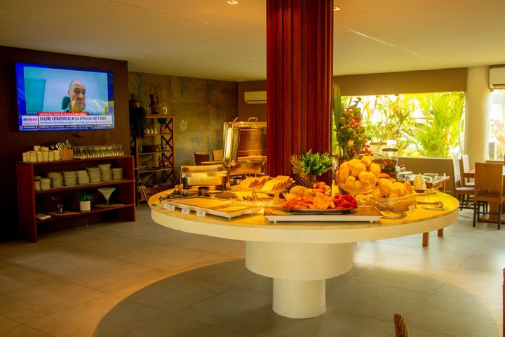 Mesa de buffet com doces e frutas em uma sala de jantar de hotel ensolarada, com uma TV ao fundo.