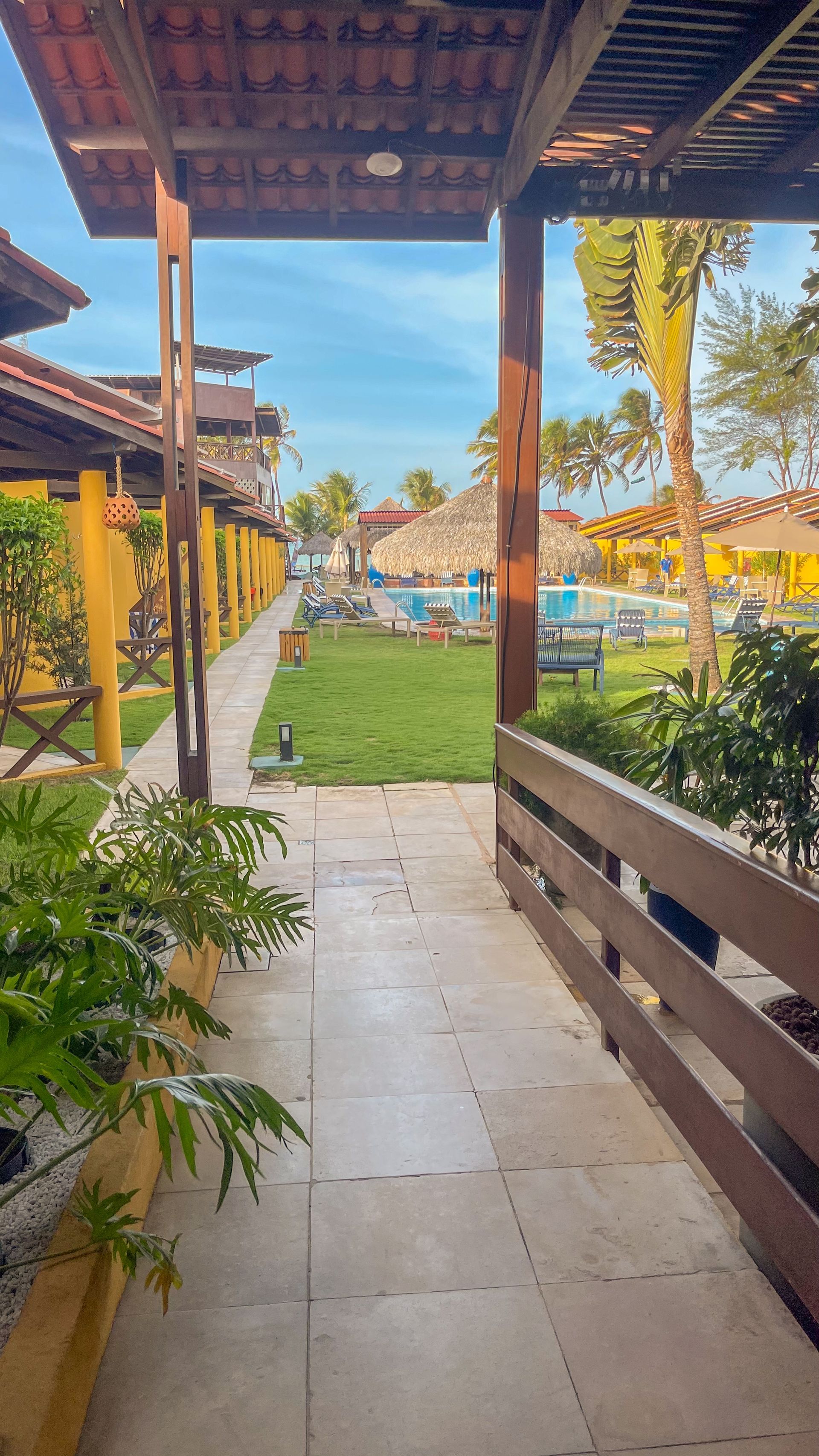 Passarela coberta no pátio que leva a um jardim ensolarado e à piscina do resort, com vigas de madeira e piso de azulejo.