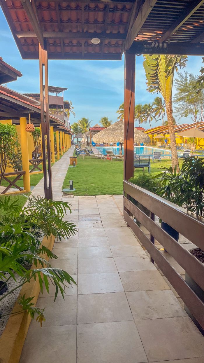 Passarela coberta que leva a um jardim ensolarado de resort com piscina, palmeiras e céu azul.