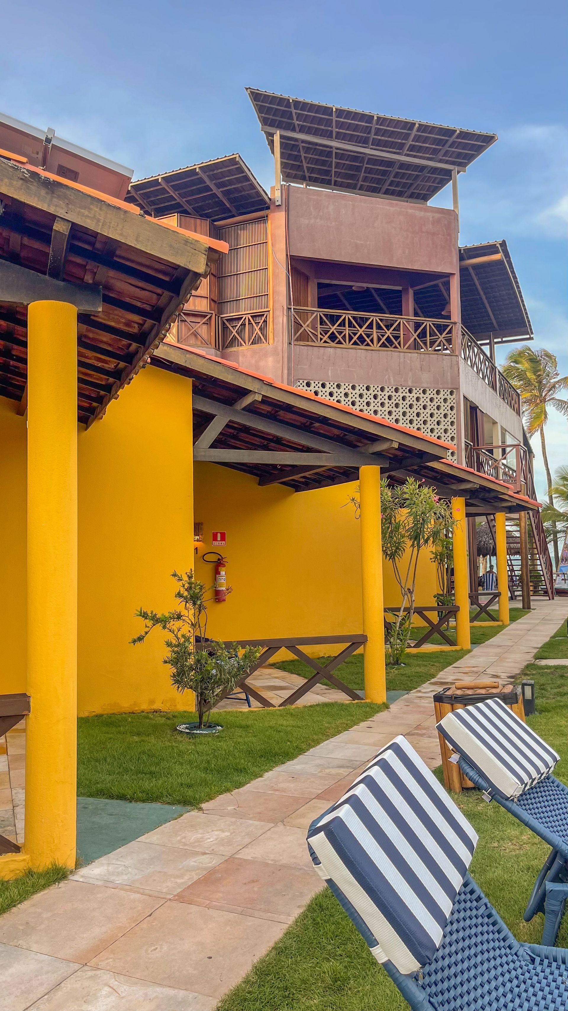 Edifícios de resort amarelo-vivo com varandas de madeira e espreguiçadeiras listradas ao longo de uma passarela.